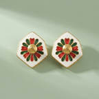 A pair of white enamel hexagonal stud earrings with a gold-tone center and red and green petal details, displayed on a soft sage green background.