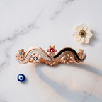 A rose gold wavy bracelet with black enamel inlays, crystal accents, and evil eye details, styled on a white marble background with a small white flower and a blue evil eye bead.