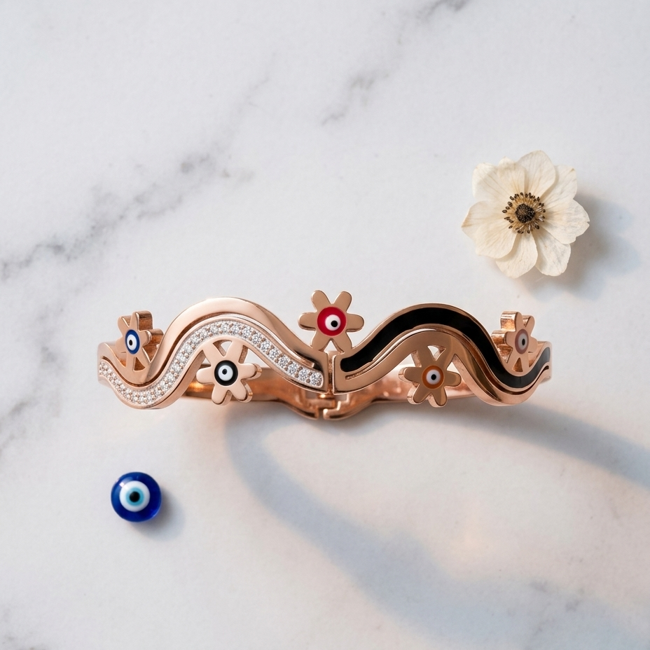 A rose gold wavy bracelet with black enamel inlays, crystal accents, and evil eye details, styled on a white marble background with a small white flower and a blue evil eye bead.
