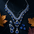 A silver-toned statement necklace with matching drop earrings, featuring deep blue teardrop and round gemstones set in silver, displayed on a dark marble surface with autumn leaves.