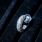 A silver crescent-shaped hoop earring paved with clear stones, displayed on a black velvet jewelry box.