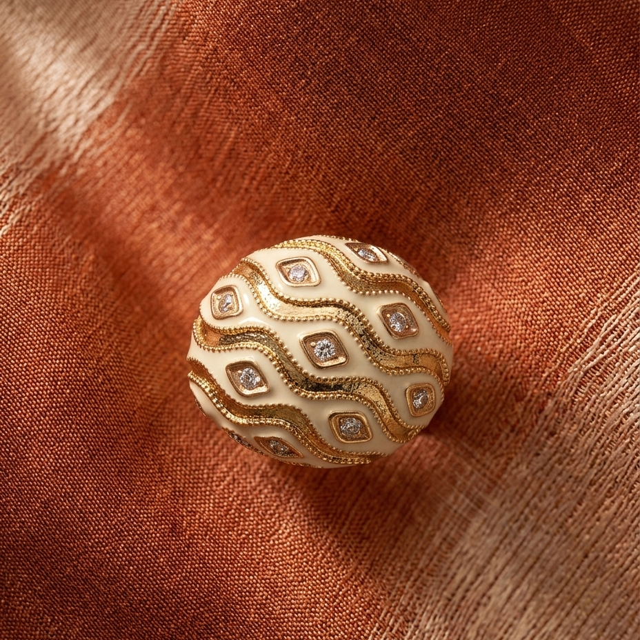 A spherical jewelry piece with a cream enamel body and gold wavy border, accented with small clear stones, resting on a textured rust-orange fabric background.