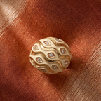 A spherical jewelry piece with a cream enamel body and gold wavy border, accented with small clear stones, resting on a textured rust-orange fabric background.