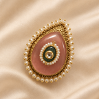 A teardrop-shaped brooch featuring a pink stone center, surrounded by a border of white pearls and gold beads, with a green circular accent and a central clear stone, displayed on a beige satin fabric background.