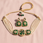 A traditional Indian jewelry set featuring a gold-plated necklace with white pearl strands, green enamel panels, and a matching pair of drop earrings, displayed on a soft pink fabric background.