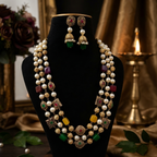 A traditional Indian jewelry set featuring a multi-strand pearl necklace with gold-plated borders, green and red stone accents, and matching gold-plated jhumka earrings, displayed on a black velvet mannequin bust.