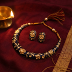 A traditional Indian jewelry set featuring a navy blue and gold necklace with floral engravings, pearl-like accents, and a matching pair of earrings, displayed on a deep red fabric next to a brass lamp.
