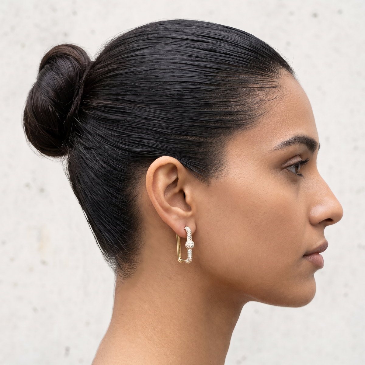 A woman in profile wearing a small gold geometric hoop earring with a white pearl-like center bead, against a light textured wall.