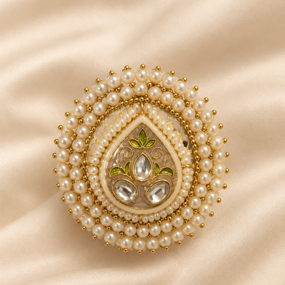 An ornate Indian-style teardrop brooch featuring a gold-plated base, surrounded by rows of white faux pearls, with green enamel floral details and clear stones, set against a draped beige satin background.