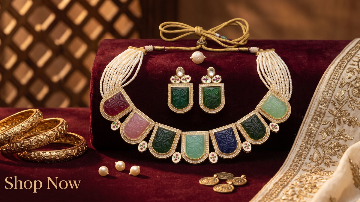 An ornate gold-toned statement necklace with matching earrings is displayed on a dark red velvet cushion, alongside stacked gold bangles, small pearl beads, and a cream embroidered dupatta. The necklace features multiple rectangular inlay panels in green, blue, pink, and purple.