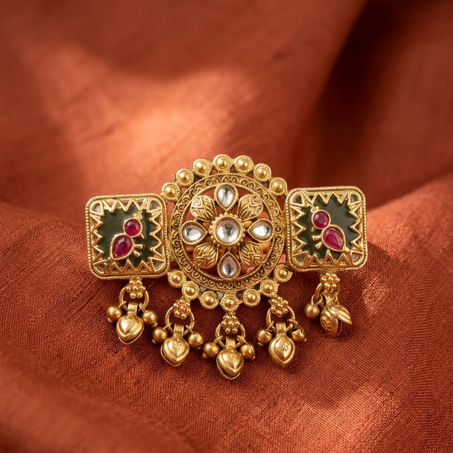 An ornate gold-toned traditional Indian brooch or belt buckle with green enamel, pink stone accents, and dangling gold bells, displayed on a draped rust-orange fabric background.