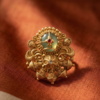 An ornate gold ring with a central oval light blue-green stone and intricate filigree details, resting on a textured rust-orange fabric background.