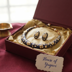 An ornate jewelry set with a necklace and matching earrings displayed inside an open burgundy presentation box lined with beige tissue paper. The jewelry features deep blue enamel, gold-toned metalwork, and small white bead accents.