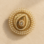 An ornate round brooch featuring a central teardrop-shaped brown stone surrounded by white faux pearls and gold-toned metal detailing, set against a textured beige background.