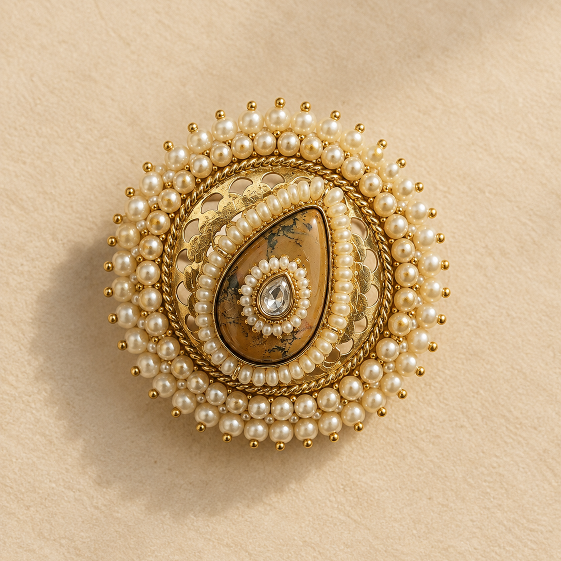 An ornate round brooch featuring a central teardrop-shaped brown stone surrounded by white faux pearls and gold-toned metal detailing, set against a textured beige background.