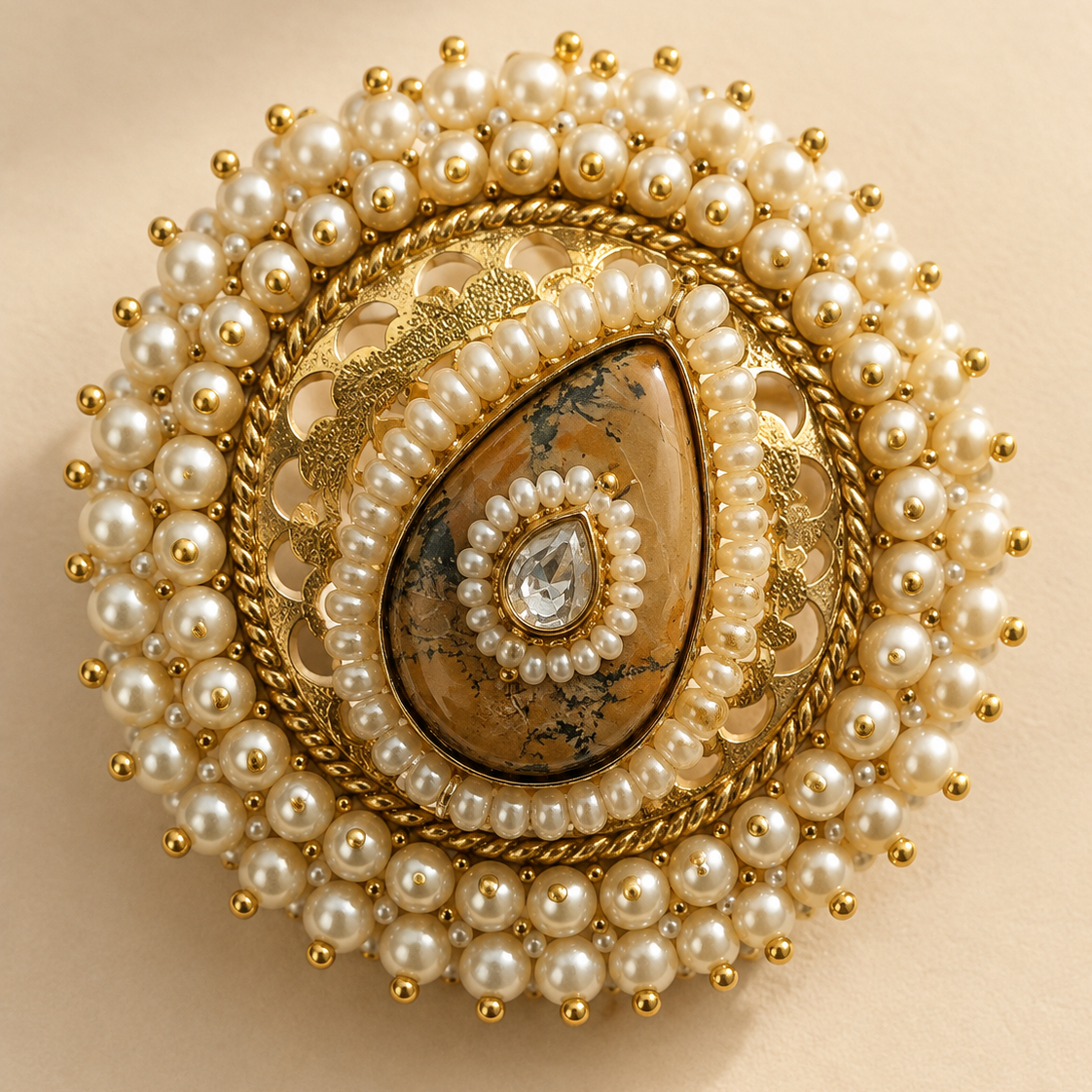 An ornate round brooch featuring a gold-toned filigree base, a central teardrop-shaped brown stone with black veining, a halo of white faux pearls, and a small clear teardrop gemstone at the center, set against a warm beige background.