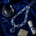 An ornate silver-toned necklace with large deep blue gemstones and clear accent stones, arranged on a dark blue velvet surface with a blue candle, loose blue stones, a rolled parchment, and a gold square pendant nearby.