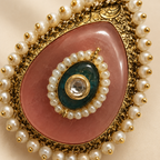 An ornate teardrop-shaped brooch featuring a large pink stone, a central dark green oval stone, a clear round gemstone, and a border of white pearls with gold accents.