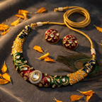 An ornate traditional necklace and matching earrings laid on a dark textured fabric with scattered orange petals and a peacock feather. The jewelry has gold-toned metal, deep red and green enamel sections, white stone accents, and a central oval stone.