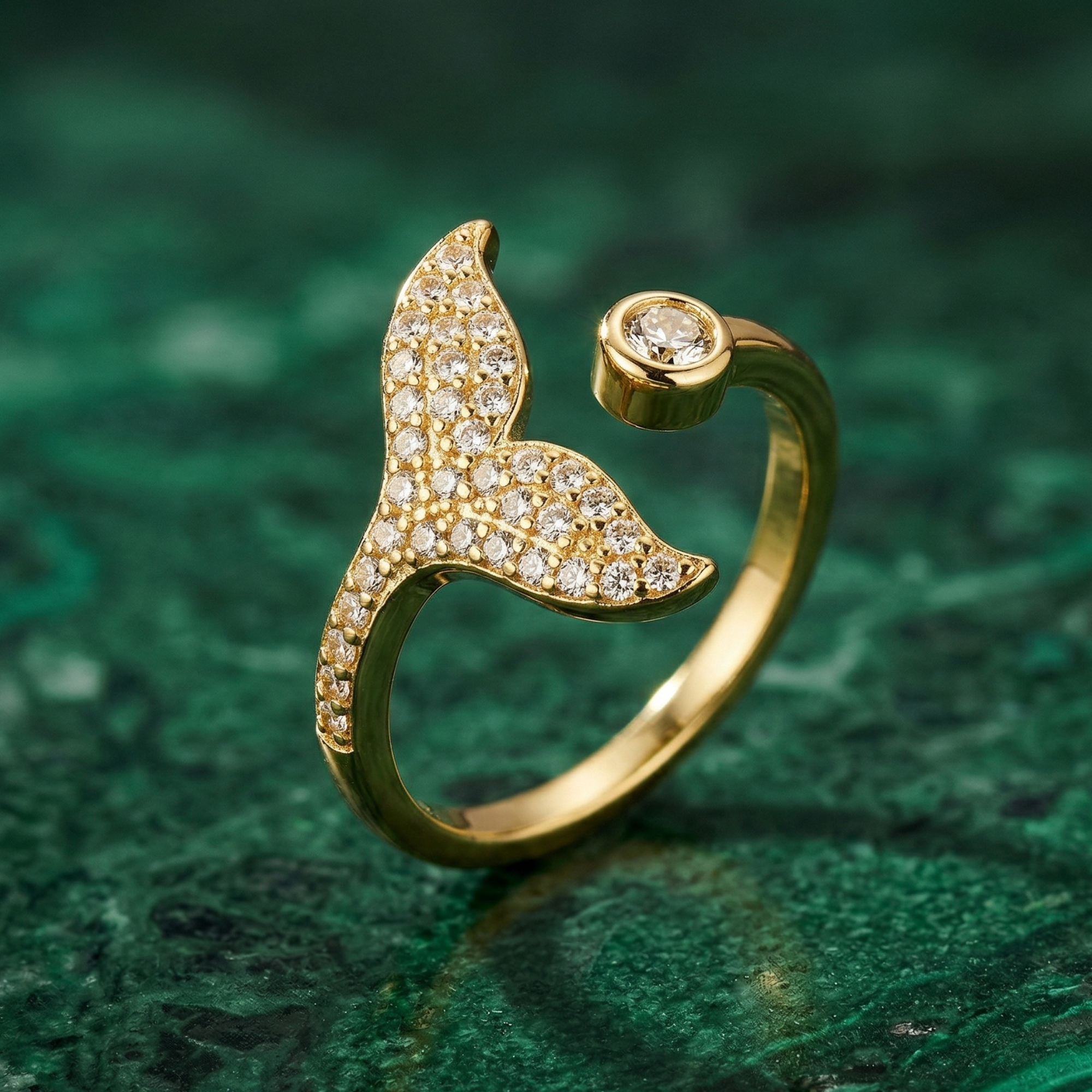 Close-up lifestyle photograph of a gold pavé mermaid tail ring worn on the ring finger or middle finger of a woman with warm olive skin, hand relaxed and slightly tilted to show the sparkling tail. Nails are neutral nude or deep emerald green. She wears a deep forest green silk sleeve or ivory cotton. Soft warm natural window light, editorial fashion photography, shallow depth of field, delicate jewellery styling, luxury brand aesthetic. Background: softly blurred warm neutral or deep green.