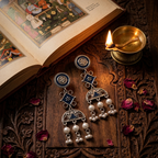 A pair of ornate silver-toned dangle earrings with blue enamel details and white pearl-like drops, displayed on a carved wooden surface next to an open book and a lit brass oil lamp.