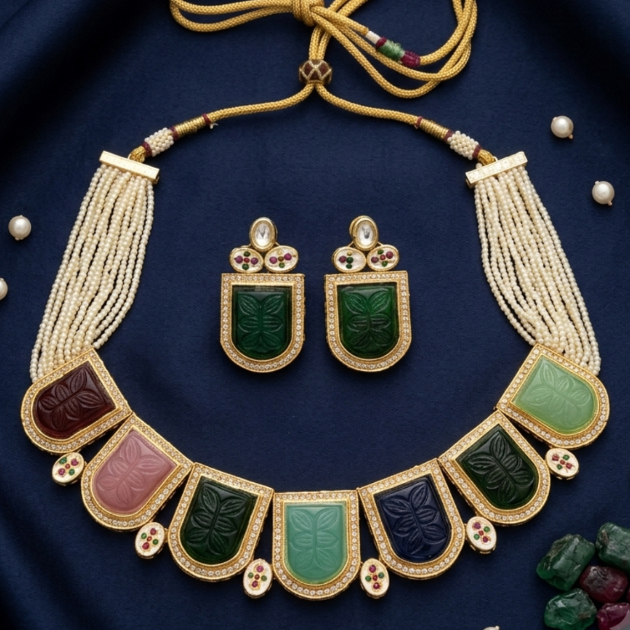 An ornate gold-toned traditional necklace set with matching rectangular earrings, displayed on a dark navy fabric background. The necklace features multiple rectangular stone panels in green, pink, and dark blue, with cream beaded fringe and a gold cord tie.