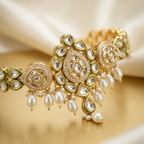An ornate gold-toned traditional Indian jewelry piece featuring white pearls, clear stones, and green enamel accents, displayed on a draped cream satin fabric.
