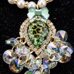 A close-up of a statement necklace featuring a large green leopard-print teardrop crystal pendant, accented with a light green bow, clear rhinestone halo, and cascading iridescent crystal clusters on a black textured background.