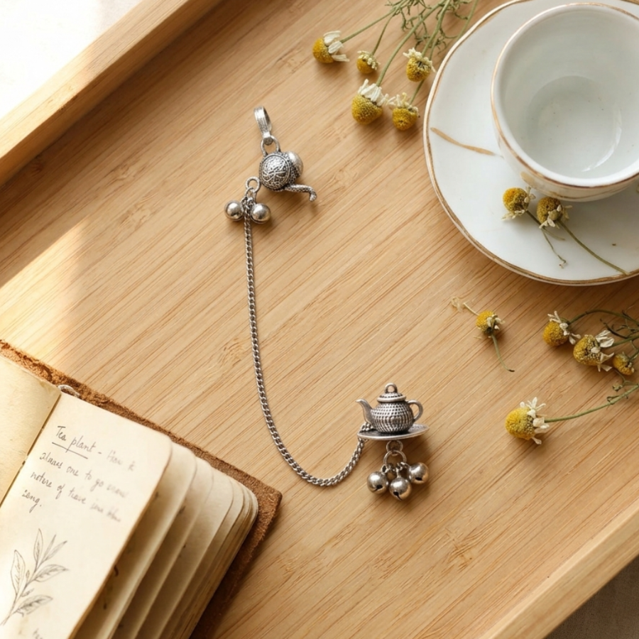 Silver bracelet with teapot charm on a wooden surface with a cup, saucer, and dried flowers.