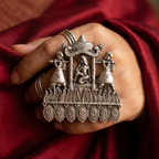 Silver decorative ring with intricate design held by a hand against a red fabric background