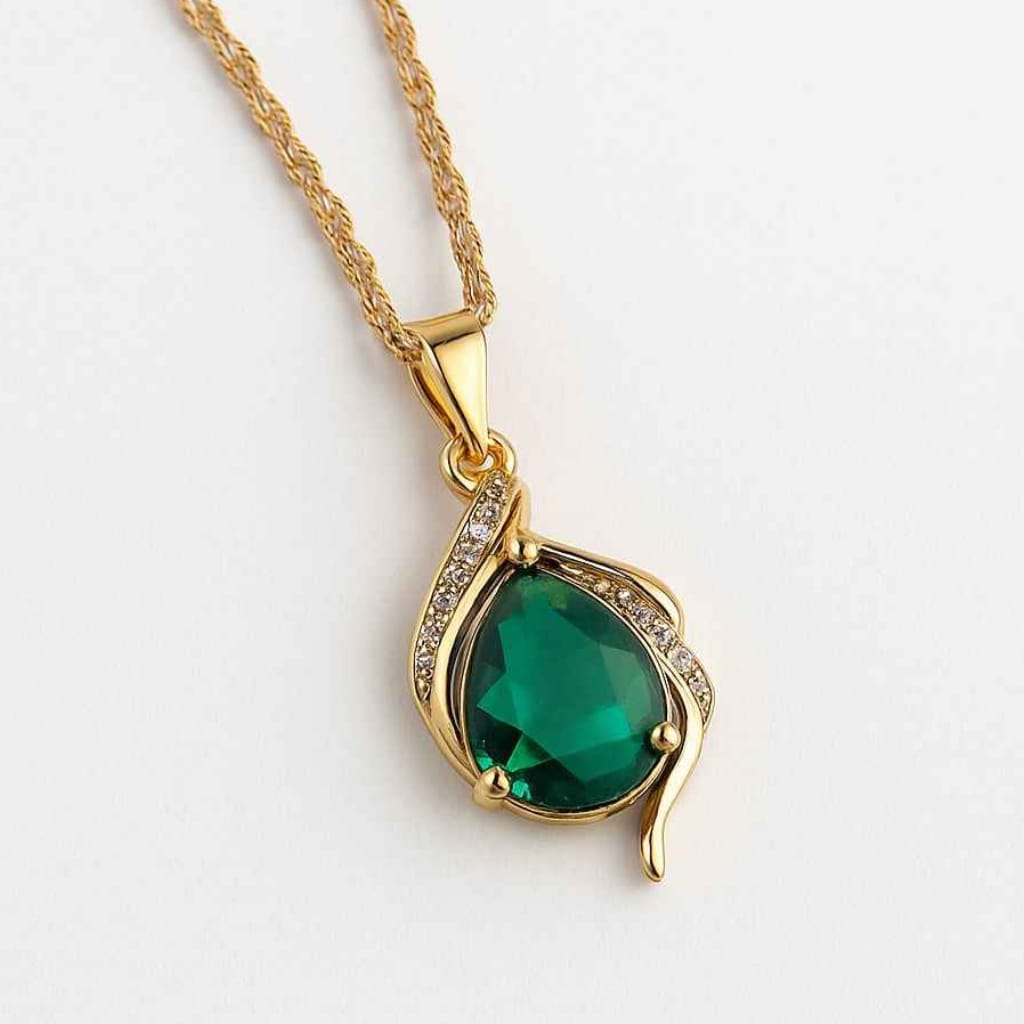 Close-up of Yugavi Tarang pendant — large multifaceted emerald green teardrop stone catching light in asymmetric gold prong setting with diagonal row of pavé white CZ crystals across wave-form gold frame, contemporary Indian jewellery