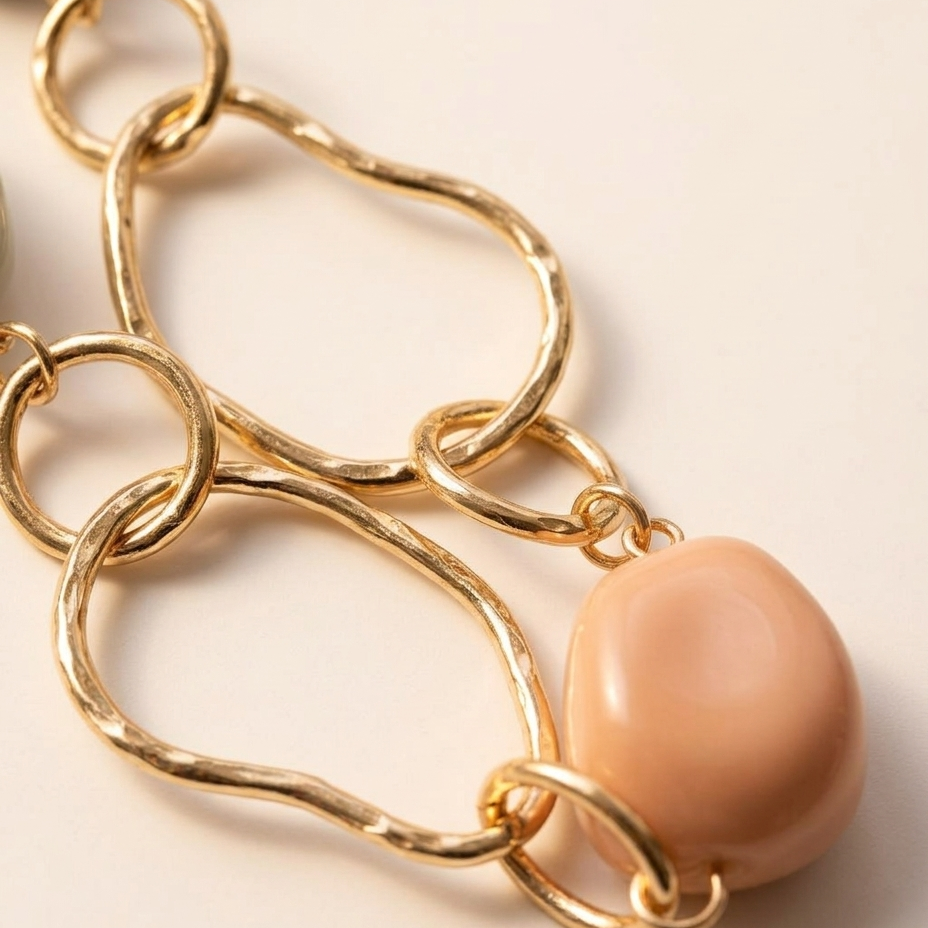 Close-up of a gold-toned chain necklace with irregular wavy links and a smooth peach-beige oval pendant on a light beige background.