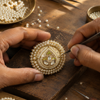 Close-up of a jeweler's hands crafting an ornate Indian-style pearl and gold pendant on a wooden workbench, surrounded by tools and loose pearls.