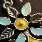 Close-up of a silver-toned floral pendant or earring charm with light blue enamel sections and a bright yellow center, photographed against a dark background.