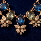 Detail of Yugavi Kshanika central sapphire stone — large deep blue marquise faceted crystal at the bottom point of the V-composition, rose gold bezel setting, pavé flower cluster beside it with champagne crystal petals catching light