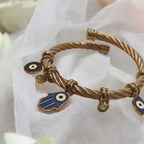 Detail of Yugavi Sharvi evil eye charms — round navy blue enamel evil eye nazar disc and elongated almond-shaped evil eye charm flanking the central Hamsa, with olive green crystal drops between them, antique gold twisted cable bangle, spiritual charm bracelet India