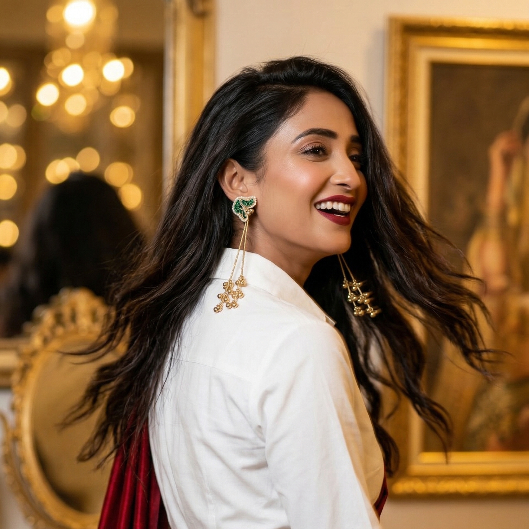Yugavi Jhankaar Emerald long drop earrings in antique gold — seed pearl-bordered peacock stud set entirely in emerald green teardrop stones, three graduated gold snake chains with clusters of tiny gold ghungroo bells, displayed as a pair on khaki fabric
