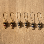 Five matching antique-style metal pendant earrings with loop tops and dangling clover-shaped charms, arranged in a row on a textured beige fabric background.