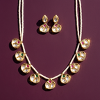 Gold-plated Kalka jewelry set featuring a necklace and matching earrings with clear stones, pink and green accent stones, and white bead strands, arranged on a deep purple background.