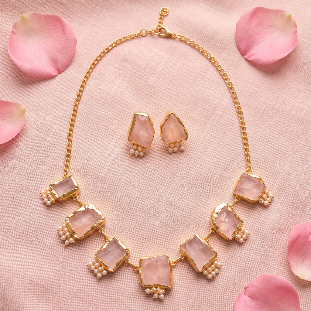 Gold-tone necklace and matching stud earrings with irregular pale pink stone centers and small pearl-like bead accents, arranged on a pink fabric background with scattered pink petals.