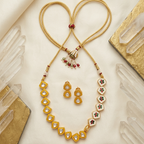 Gold-toned jewelry set laid flat on a cream fabric background, including a long beaded necklace with a heart-shaped loop at the top, matching gold earrings, and decorative crystal props with gold and wood accents around the edges.
