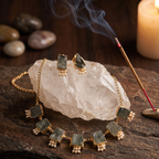 Gold-toned jewelry set with rectangular dark green stones and small white bead drops, displayed on a clear crystal cluster over a rustic wooden surface, with a burning incense stick and a lit candle in the background.