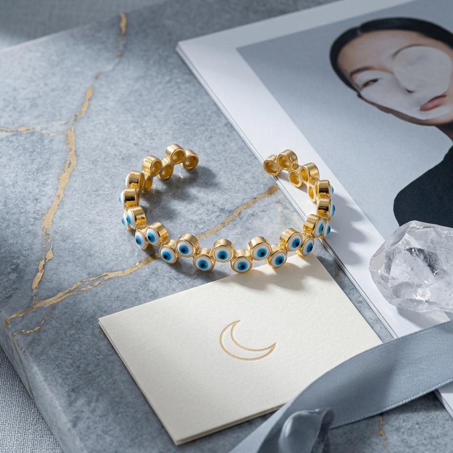 Gold cuff bracelet with blue and white evil eye beads, displayed on a gray marble surface with a cream card, satin ribbon, and magazine page.