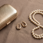 Gold hoop earrings displayed on a textured taupe surface next to a gold case and a pearl necklace.
