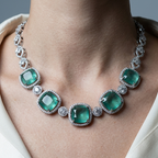 A close-up of a model wearing a silver-toned necklace featuring seven large emerald-cut green gemstones and small clear accent stones, styled with a cream-colored blouse.
