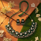 A green and silver traditional Indian jewelry set featuring a necklace and matching earrings, styled on a terracotta tile and banana leaf with dried white flowers.
