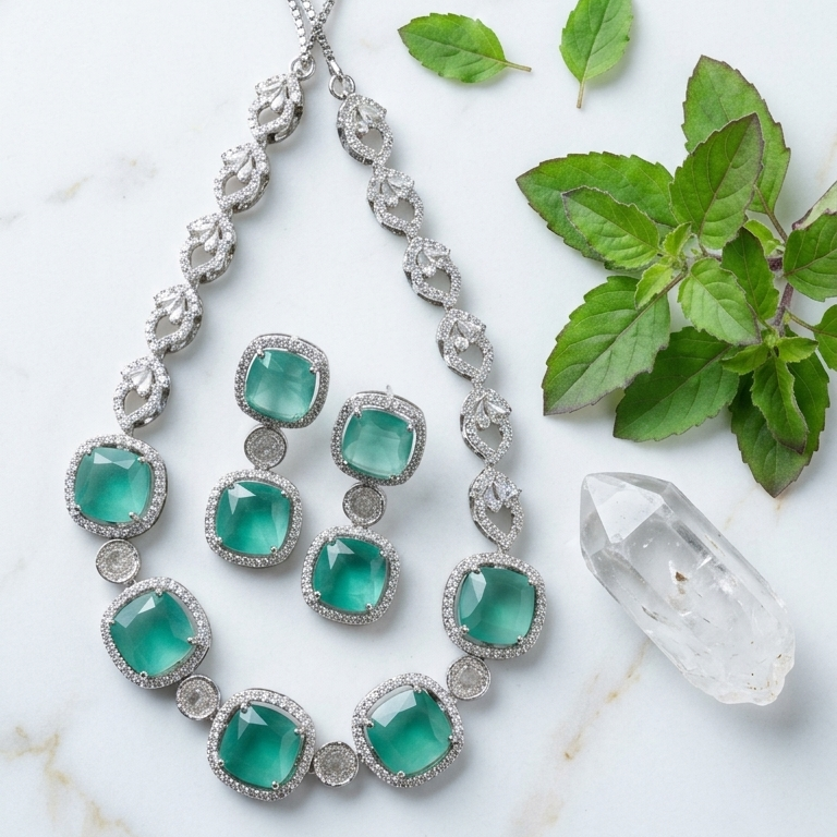 Silver-toned jewelry set with emerald-cut green stones, including a teardrop pendant necklace, matching drop earrings, and small stud earrings, arranged on a white marble surface with green leaves and a clear crystal.