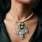A close-up of a model wearing a pearl necklace with a large, ornate pendant featuring a green marquise-cut center stone and iridescent crystal clusters, styled with a dark green satin top.