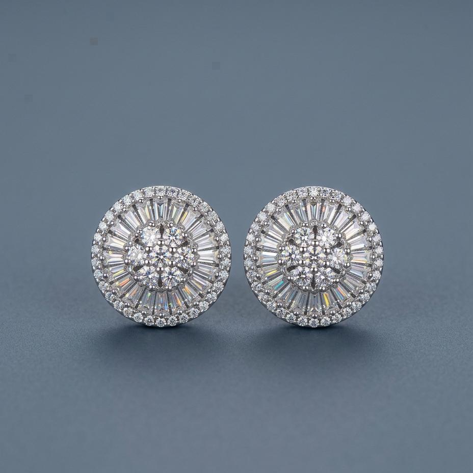 A pair of round silver-toned stud earrings with intricate halo and baguette crystal detailing, displayed on a dark slate gray background.