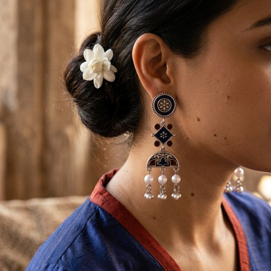 A close-up of a woman wearing a traditional Indian jhumka earring. The earring features a silver-toned metal frame with black enamel detailing, a central geometric motif, and three dangling pearl-like beads.